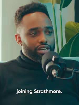 A man with short, dark hair and a trimmed beard is speaking into a microphone, likely in an interview setting. He's wearing a black turtleneck sweater. The background includes a soft-focus plant. The lighting is warm and inviting.