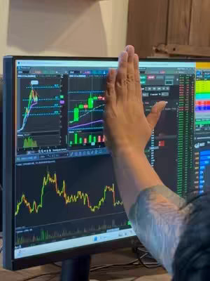 A person's hand is held up in front of a computer screen displaying stock market graphs. The hand is positioned to the mid-right of the screen, partially obscuring the data. The screen itself is a monitor showing detailed stock charts with green and red candlesticks, lines, and data points. The background visible in the image includes a wooden panel, adding a neutral tone to the scene.