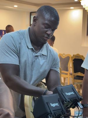 A man with a shaved head is looking down while holding two video cameras. He is wearing a gray polo shirt. Behind him, there are hints of an interior, including ornate chairs and other blurred figures.