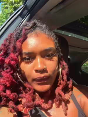 A woman with red-toned dreadlocks sits in the driver's seat of a car, with sunlight streaming in. She has a nose ring and hoop earrings. The image includes parts of the car interior and a view of the outdoors visible through the windows.