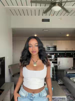 A young woman with long, wavy dark hair is posing in a well-lit room. She's wearing a white crop top and light-wash jeans, and a necklace with a round pendant. The background includes a ceiling with slats, a ceiling fan, and a glimpse of a kitchen area.