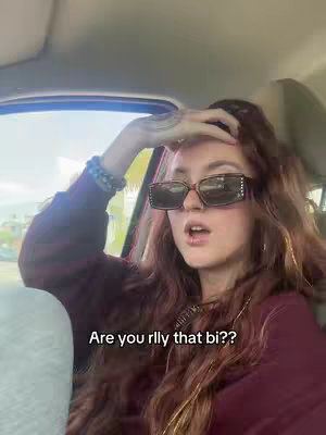 A young woman with long, wavy red hair and sunglasses is seated inside a car. The text overlay says: Are you rlly that bi??