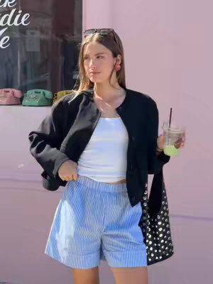 A woman stands in front of a pink wall with sunglasses on her head, wearing a black jacket, a white top, and striped blue shorts. She holds a drink in one hand and a bag in the other.