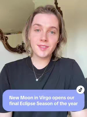 A person with light hair and fair skin is centered with a friendly expression and slight smile. They wear a black t-shirt and a delicate silver necklace. A blue rectangular text overlay at the bottom of the frame, labeled "New Moon in Virgo opens our final Eclipse Season of the year". The background is a neutral-toned room with a decorative mirror.
