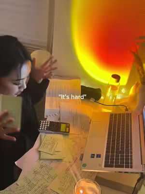 A woman stands to the left, reaching towards a chaotic desk. Papers and a calculator are visible. A laptop sits on the right, and a sunset lamp casts an orange glow.