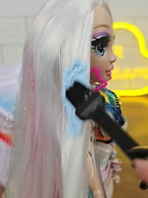 The image shows a close-up of a Rainbow High doll's face in profile, with its long white hair being worked on with a tool, likely a styling brush. The doll has detailed facial features, including bright eye makeup and a painted face. Behind the doll, the background suggests a brightly lit environment, possibly a salon setup; a large neon yellow shape adds visual interest.
