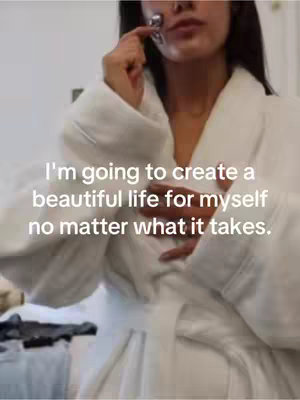 The image shows a woman in a white bathrobe, partially visible from the chest up, in a room with a simple, light-colored background. She is holding a facial massage tool to her cheek and appears to be looking down. Text overlayed on the image says, 'I'm going to create a beautiful life for myself, no matter what it takes.' The composition is simple, with the focus on the woman and the message.