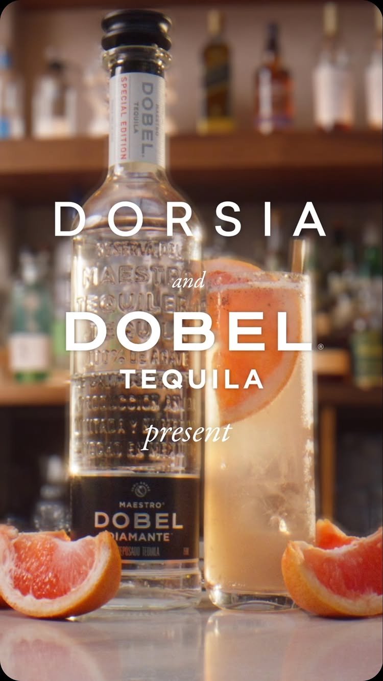 The image showcases a well-lit bar scene with a focus on a tequila bottle, a cocktail, and grapefruit slices. The composition is balanced, with the Dobel tequila bottle prominently featured on the left, the refreshing Ace Paloma cocktail to its right, and grapefruit slices at the bottom. The background has a bar or restaurant setup, and the color palette is warm, with a mix of browns and bright oranges, creating an inviting feel.