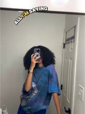 A selfie taken in a bathroom mirror. The person is holding the phone in front of their face, obscuring it. They have large, curly black hair. They wear a tie-dye blue and purple t-shirt and a watch on their left wrist. Above their head, on the mirror, is an overlaid text in a yellow bubble. The background includes a white door, a towel rack, and a part of the bathroom.