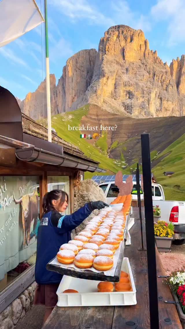 A woman is holding a tray of donuts in front of a food stand, with the Dolomites in the background. The woman is wearing a blue jacket and black gloves.