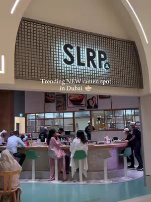 A well-lit interior shot of SLRP by 3Fils. The foreground features a bar with people seated at it, with green seats. Above, a sign that reads "SLRP &" is visible. The overall composition is a restaurant scene, showcasing a vibrant atmosphere.