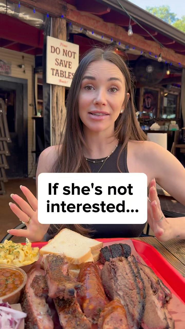 A woman with long brown hair is sitting at a table, holding her hands up. There's a plate of BBQ food in front of her, including brisket, sausage, and other items. A text overlay reads: "If she's not interested..."