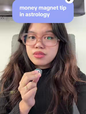 A woman with long, wavy dark hair and glasses is seated in a well-lit space, holding a small, circular object in her right hand. She has a neutral expression, and a text overlay at the top of the image reads, "money magnet tip in astrology."