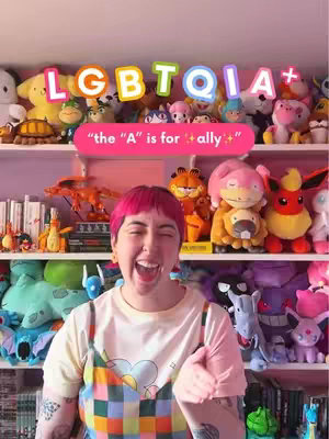 A woman with pink hair is centered, laughing widely, in front of shelves filled with plush toys and books. The word 'LGBTQIA+' is in colorful, bubbly letters above her head, with a pink banner that says 'the "A" is for ally' on in the video. The setting has a playful, vibrant atmosphere.