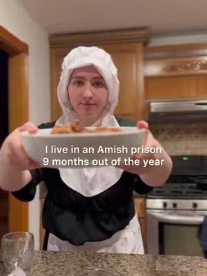 A person in a white Amish bonnet holds a plate of food dishes while standing in a kitchen. A text overlay reads, "I live in an Amish prison
9 months out of the year."