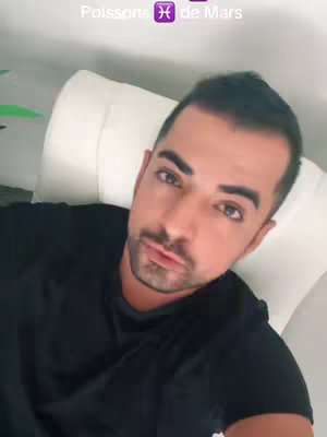 A close-up shot of a man in a black shirt leaning against a white surface. The man has short, dark hair and a trimmed beard and mustache. Above his head, text reads "Poissons" with the Pisces symbol and "de Mars". The background is out of focus and appears to be an interior setting.