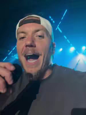 A man with short brown hair and a white cap is holding a microphone and speaking passionately while looking up, with a stage background with lighting.