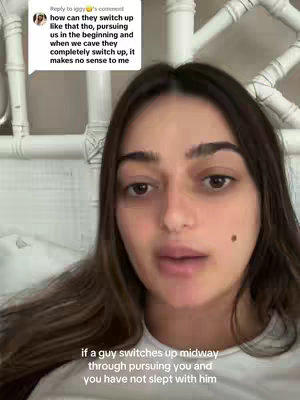 A woman with long, dark hair is centered in the frame, presenting a video. She is positioned against a backdrop showing a white, ornamented headboard and part of a wall. The video appears to be filmed indoors. The woman’s facial expression and direct gaze give a sense of her speaking to the audience. Text overlays are added to the screen, enhancing the primary content of the video.