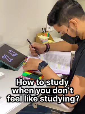 A person, wearing a black mask and watch, is sitting at a desk and studying, with a tablet and other stationary on the desk. The text overlay reads: How to study when you don’t feel like studying?