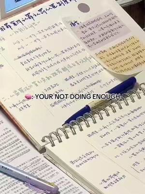 The image shows a close-up of a spiral notebook open on a desk, with other papers and a pen present. The notebook has handwritten notes in blue ink with some Chinese characters, and a blue pen is placed on top of the notebook. There's a pink sticker and a text overlay that says 'YOUR NOT DOING ENOUGH'. The background is slightly blurred, with a neutral color palette.