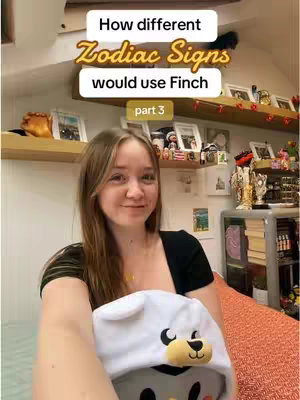 A selfie-style shot featuring a young woman holding a stuffed animal shaped like a sheep wearing a hat with the same design features. The background shows a room with shelves holding various objects, and text overlays state the video is about zodiac signs using Finch.