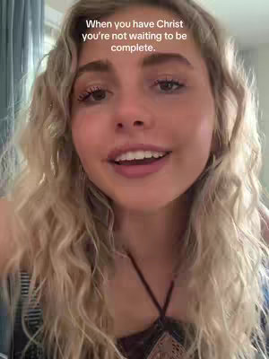A close-up shot of a woman with long, wavy blonde hair, smiling and looking directly at the camera. The background is slightly blurred, suggesting an indoor setting with possibly a window or light source to her left. She appears to be speaking.