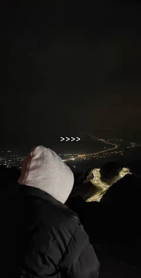 A nighttime shot overlooking a city, with a person in the foreground wearing a white hooded jacket, their back to the camera. The city lights create a sparkling backdrop in the distance, and the overall composition is dark and moody.