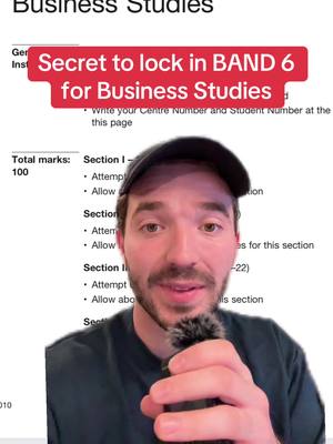 A man with a beard and a black cap holding a microphone in front of a paper with business studies topics; Hook text in a red box at the top.