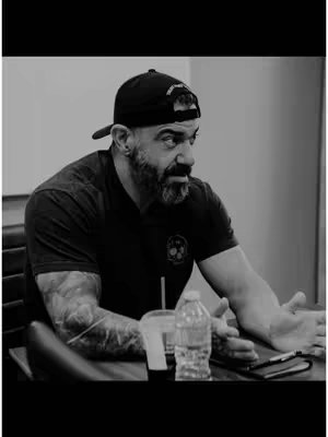 A monochrome image features a man in a black cap and shirt, seated at a table. He has a beard and tattoos on his arm. A water bottle and drink cup are visible on the table.