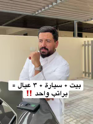 A man in traditional white attire stands outdoors, appearing to contemplate. A black and silver laptop sits in the foreground, with the man's phone on top. The background features a modern architectural style with a white wall and a slatted roof structure.