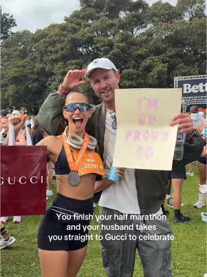 A photo of a woman and a man celebrating a half marathon finish. The woman is smiling with sunglasses on, holding a Gucci shopping bag and wearing a medal. The man is wearing a white cap and holding a sign that says, I'M SO PROUD SO SO. Behind them is a park setting with large trees.