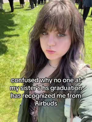 A selfie-style shot featuring a young woman with long hair and a neutral expression. She is standing in front of a green, grassy background with several people visible in the distance, suggesting an outdoor event, like a graduation. Overlayed text at the bottom declares that she is confused because nobody recognizes her from "Airbuds".