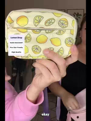 A person's hands are holding a small, patterned wallet with lemon designs against a neutral background. To the right, another hand holds a pink purse, with parts of the person's arm and chest visible, wearing a black top. A sign is attached to the wallet.