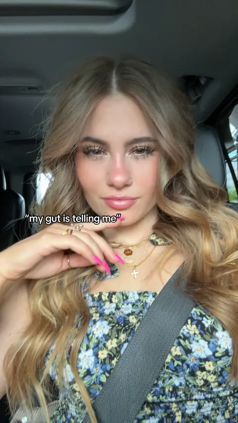 A selfie taken from inside a car. The primary figure is a young woman with long, wavy blonde hair. She has full lips, pink nail polish, and multiple necklaces, including a cross. She's wearing a floral patterned top, with a seatbelt across her chest. The background shows the interior of a car with visible headrests and a glimpse of the outside environment.
