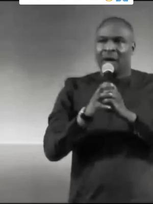 A grayscale image shows a man speaking into a microphone. He is wearing a dark-colored shirt, and his hands are clasped in front of the microphone. The background is a plain, light-colored wall.