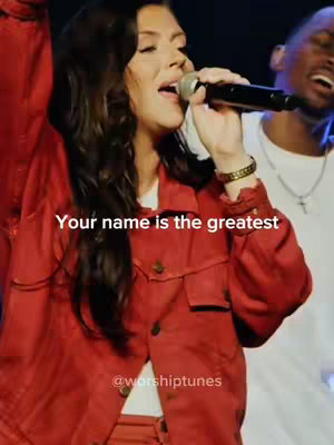 A woman with long, dark hair sings into a microphone. She wears a red jacket over a white shirt. The background is dark with other figures present.