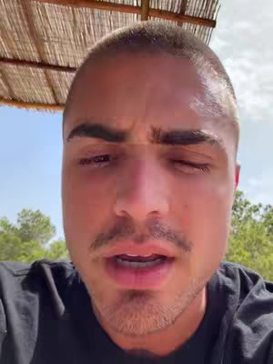 The image shows a close-up of a man's face. He has short, shaved hair and a mustache and goatee. He appears to be speaking, with his mouth slightly open. The background includes a blurred view of vegetation and a light-colored, woven canopy overhead.