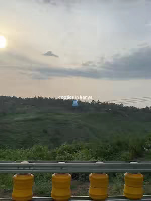 The cover image presents a wide outdoor shot taken during the day, the sun is at the top left and is creating a lot of light. A road barrier in the foreground is constructed with yellow pillars and chrome rails, while a field of green foliage stretches up to a distant rise covered in trees. Above, the sky is a display of clouds.