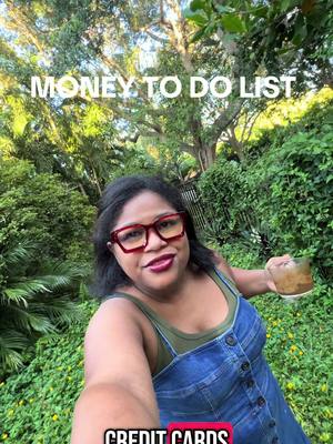 A selfie-style shot with a woman in a garden setting. The woman wears a denim dress, red-framed glasses, and holds a drink. Overlaid text at the top says "MONEY TO DO LIST" and text at the bottom says "CREDIT CARDS."