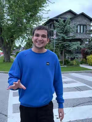 A man wearing a blue sweatshirt stands in front of a crosswalk. He is smiling and gesturing with his right hand. Behind him are trees, a house, and green grass as the background.