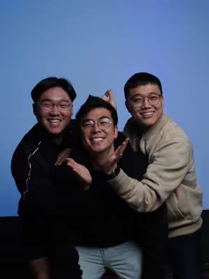 Three young men smiling and posing for a photo. They are standing in front of a blue background. The man in the center is wearing a black sweater and is being embraced by the two men on either side of him, who are also wearing glasses. The man on the right is wearing a beige jacket. All three are looking at the camera and seem to be in a joyful mood.