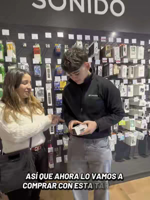 A woman and a man stand inside a store, possibly an electronics shop, in front of a wall displaying various product packages. The woman is on the left, pointing, while the man, holding a small item, looks down. Below them, text overlays.
