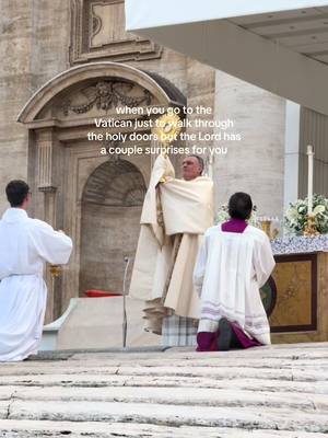 The image shows a religious ceremony at the Vatican. A priest, dressed in white and holding a golden monstrance, is the central figure. Two other figures, also in white robes, are kneeling. Behind them is an archway on a stone building.