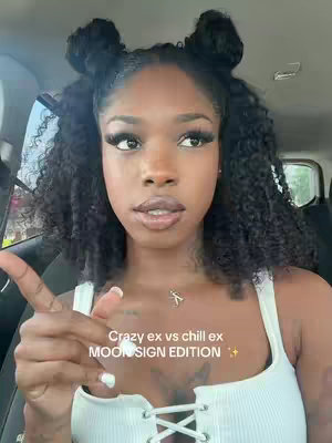 A young woman with dark curly hair styled in two buns looks toward the camera, slightly pointing with the index finger of her left hand. The background is blurred, suggesting she is in a vehicle.
