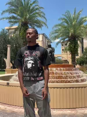 A young Black man stands in front of a multi-tiered fountain on a sunny day. He wears a black t-shirt with a graphic and gray athletic pants, with a backpack on. Behind him are palm trees and buildings, with a statue on top of the fountain, composing a bright daytime scene.