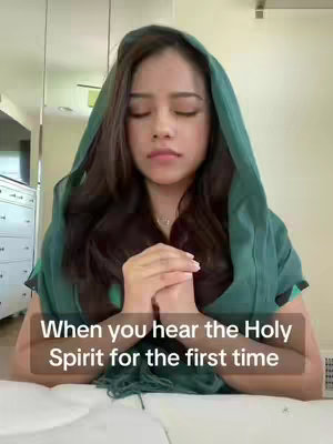 A woman with long dark hair, wearing a green hood, is depicted with her hands clasped in front of her. She has her eyes closed expression as praying. The background is blurred but seems to be an interior space. The text overlay is in a dark-colored rectangular box at the bottom, containing white text.