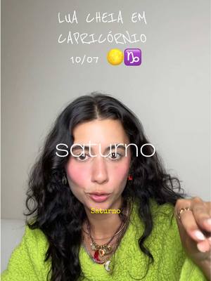 A selfie-style shot with a woman in the center. The background is a simple, light gray wall. The woman has long, dark, wavy hair. She is wearing a green textured jacket. Her cheeks are flushed with pink blush. Above her head is white script that reads 'LUA CHEIA EM CAPRICÓRNIO' (Full Moon in Capricorn). Below that is 10/07 with a moon and Capricorn emoji. The word 'saturno' (Saturn) is written in white across the upper portion of the image and under her chin there is text that says 'Saturno'.