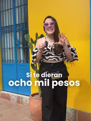 A woman stands in front of a yellow wall, holding a microphone and gesturing with her hand. The background includes a blue door and some greenery. Text overlayed on the image asks, "Si te dieran ocho mil pesos?"