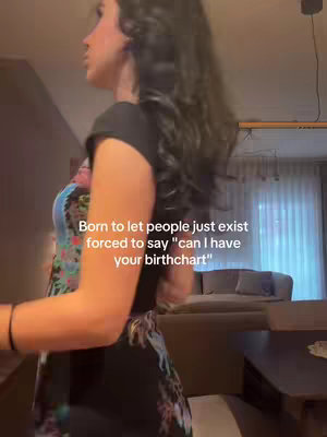 A young woman stands in a living room, angled away from the camera. She is wearing a black top and floral dress, with long black hair. Text overlayed on the image reads: Born to let people just exist forced to say "can I have your birthchart". The overall tone appears to be centered on astrology and a hint of humor.