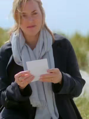 Kate Winslet is holding a white card, standing outdoors, slightly facing the camera with a blurred background of green foliage. She is wearing a black coat, a light blue scarf, and a ring on her finger. The lighting appears soft and natural.